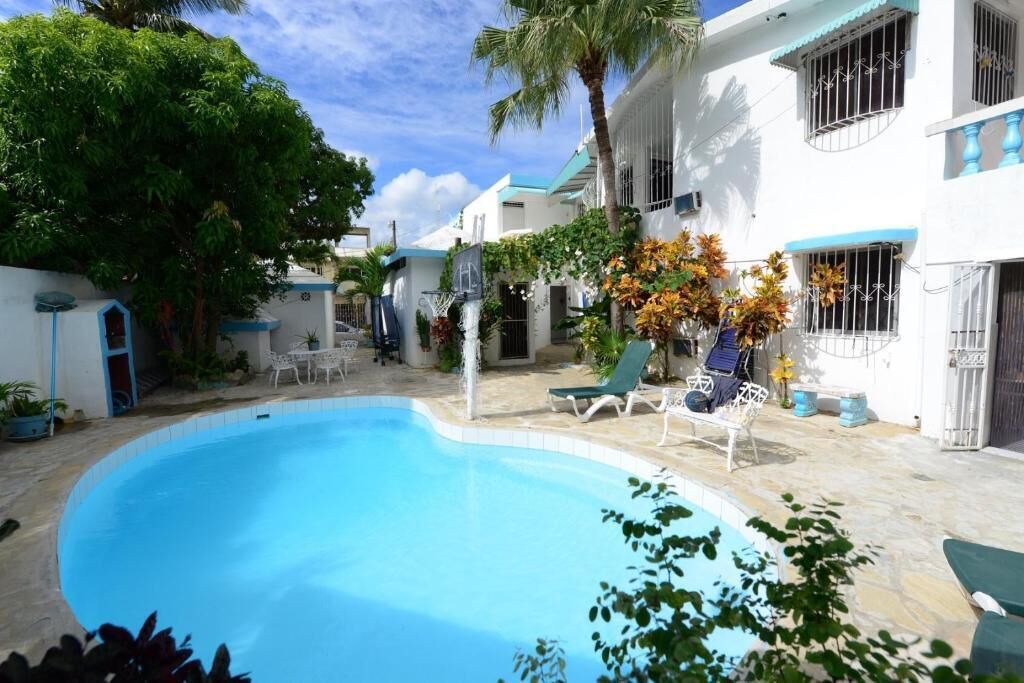 Una piscina con sillas de descanso, rodeada de edificios blancos con detalles azules y palmeras.