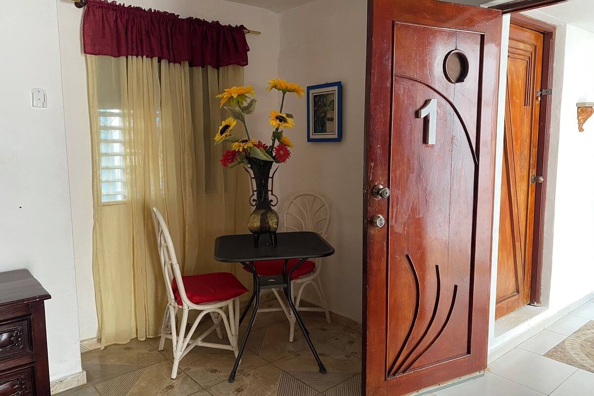 A room with a wooden door marked with the number 1, a dining table with two chairs, a vase with flowers, and a window with curtains.
