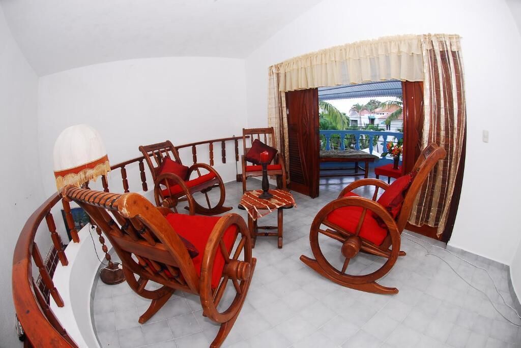 A balcony with wooden rocking chairs and a table, white walls, and a view of the outside.