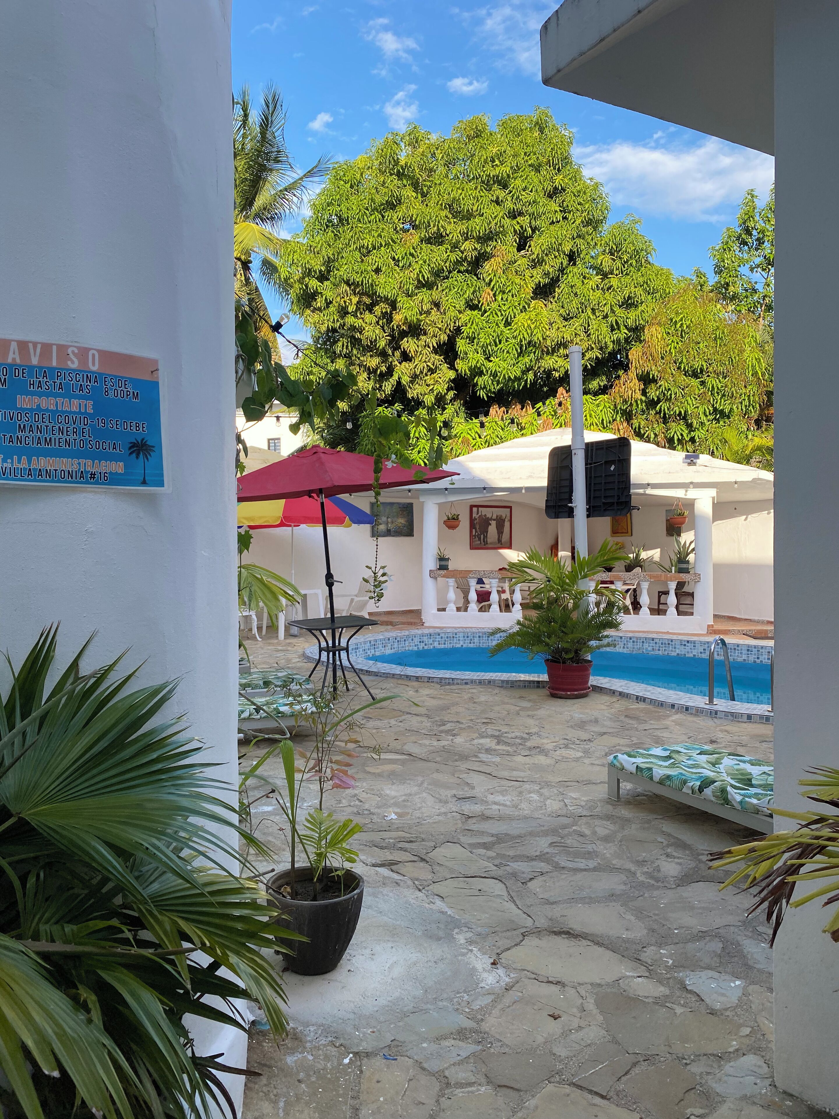 A pool area with a sign indicating closing time and COVID-19 guidelines, surrounded by potted plants and outdoor furniture.