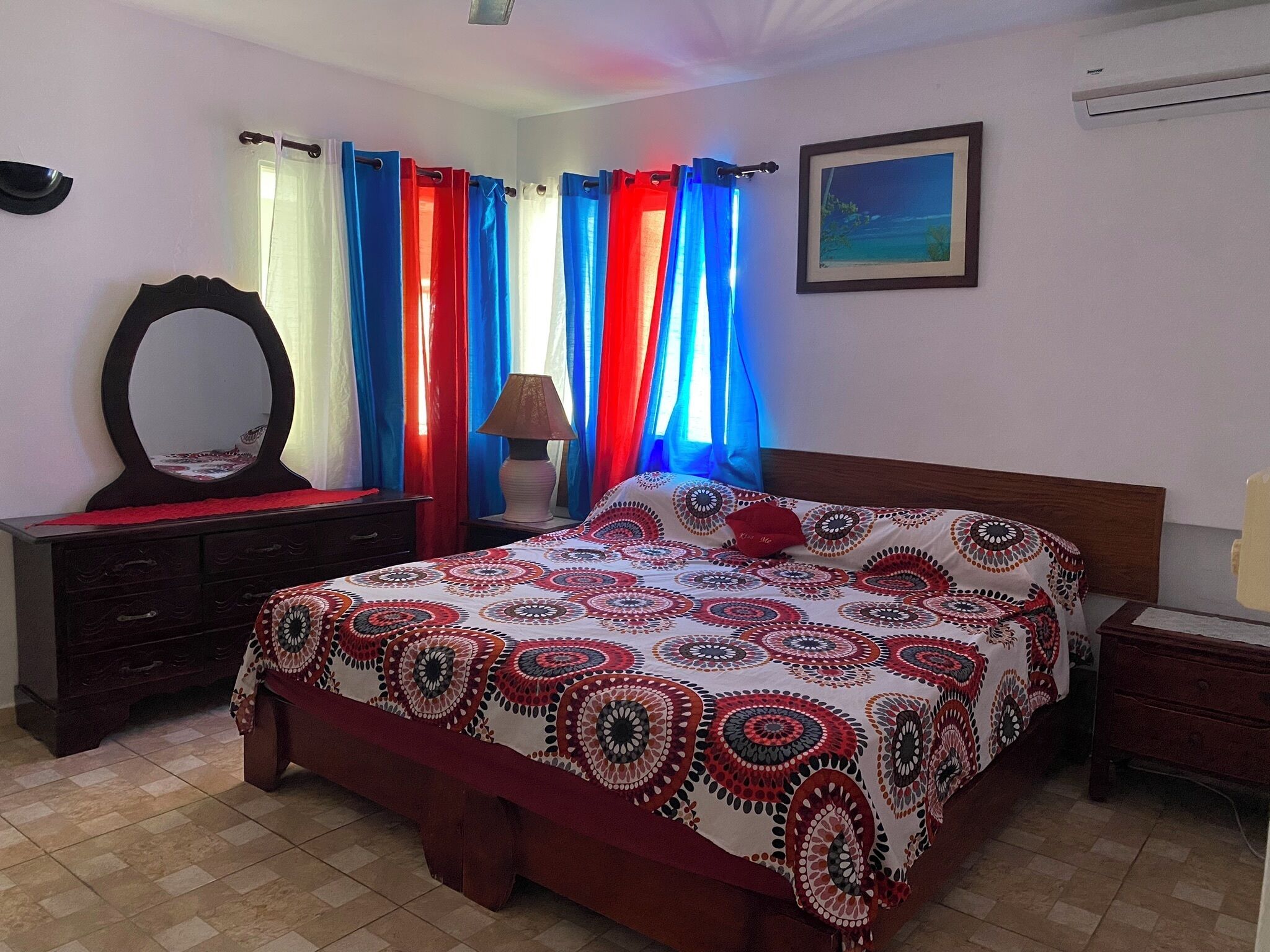 Una camera da letto con un letto, una cassettiera, uno specchio, tende, una lampada e un quadro appeso al muro.