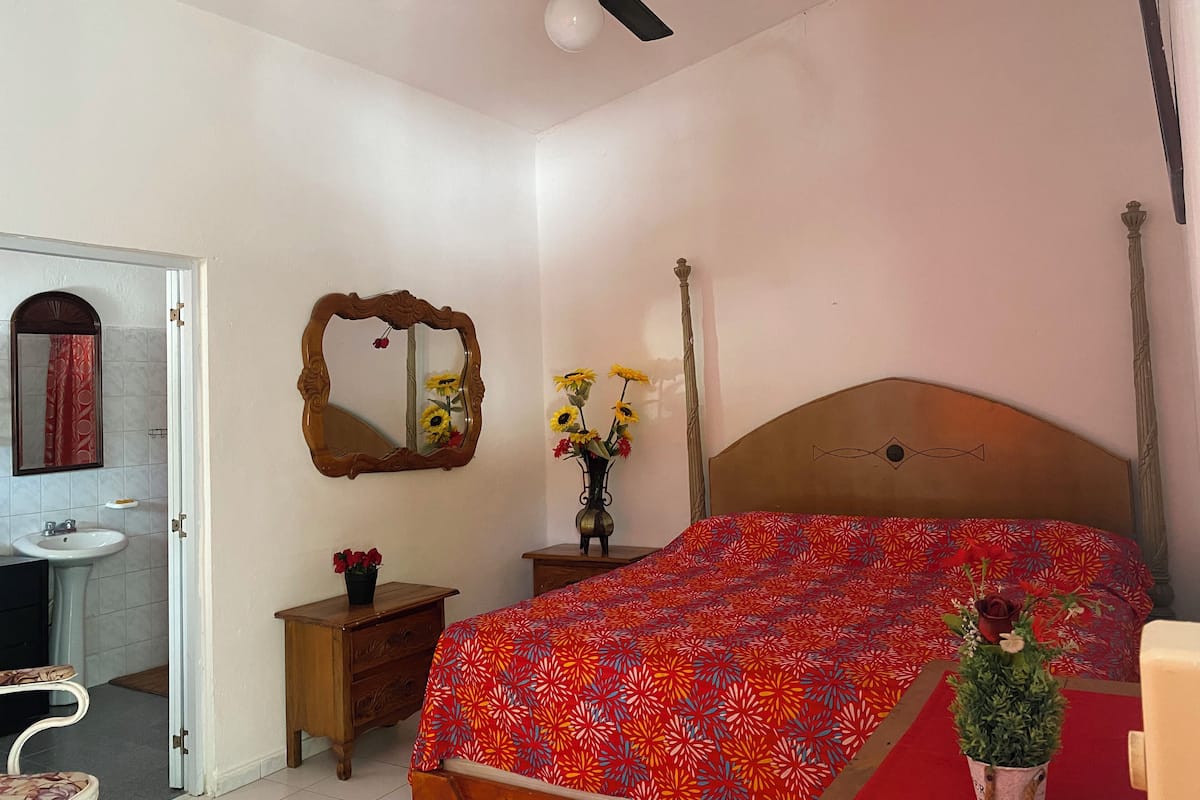 A bedroom with a bed, a bedside table with a vase of flowers, a mirror, and a ceiling fan.