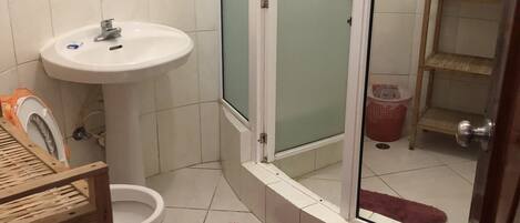 Un baño con lavamanos blanco, inodoro, ducha con puertas de vidrio satinado y estante de madera.