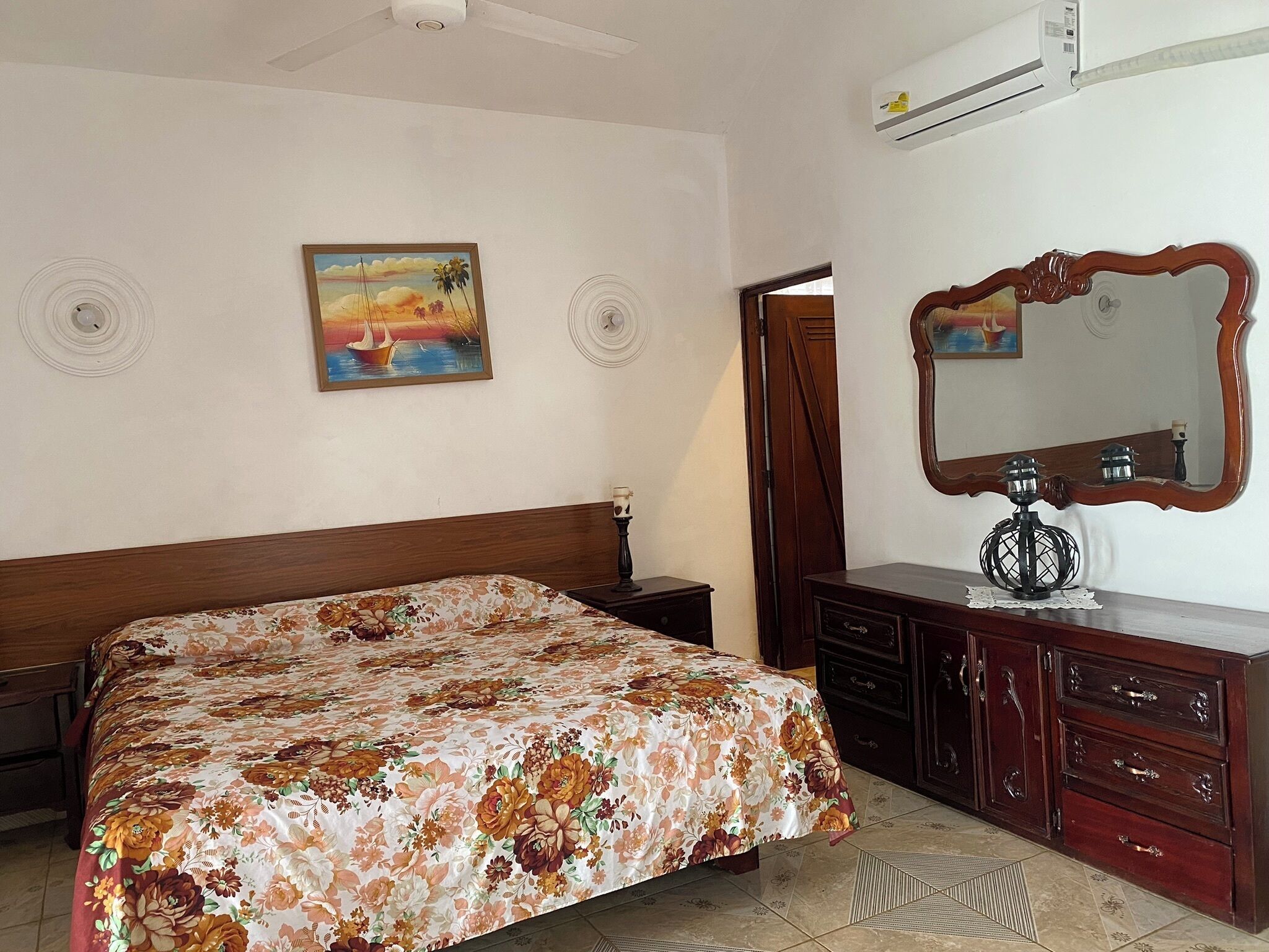 A bedroom with a floral bedspread, a wooden headboard, a dresser with drawers, a mirror, and a painting on the wall.