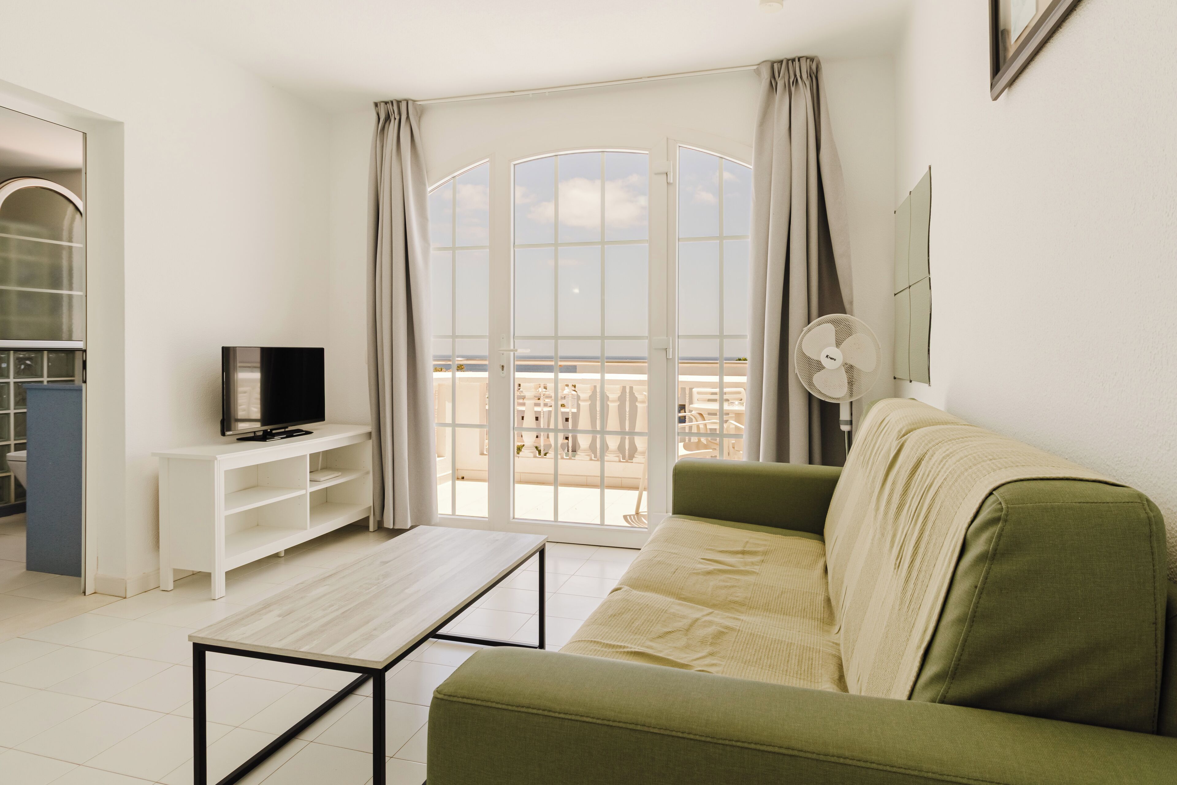 Una sala de estar con un sofá verde, un televisor sobre un mueble blanco y un balcón con vistas.