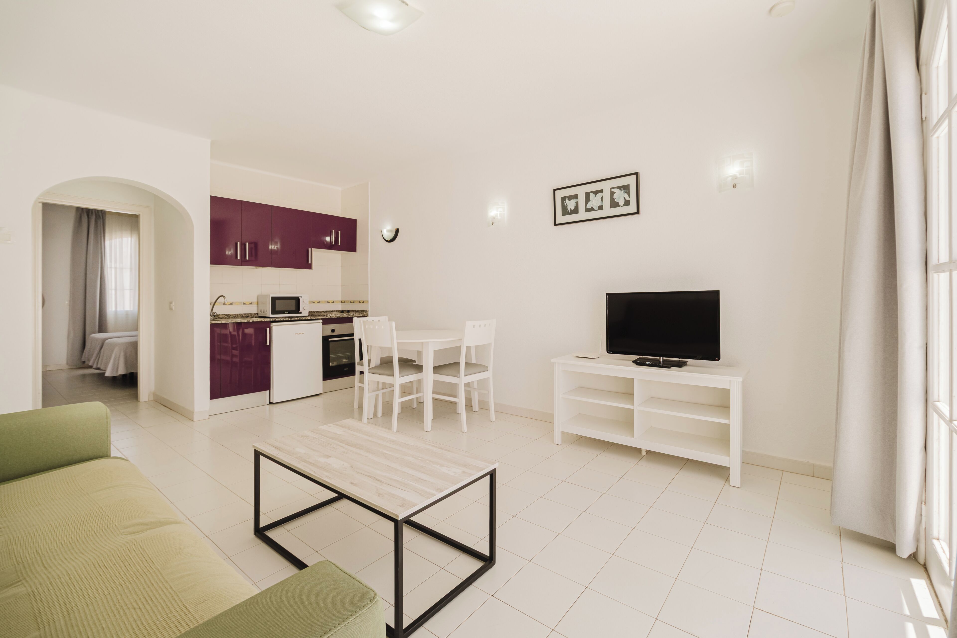 Un salon moderne avec un canapé vert, une table basse, un téléviseur posé sur un meuble blanc, un coin repas avec des chaises et une cuisine aux armoires violettes.