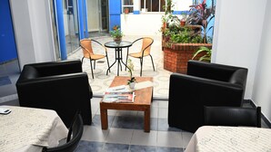 Eine Hotelhalle mit Glastür, die zu einer Terrassenfläche führt. Innen befinden sich zwei schwarze Ledersofas, ein Holztisch mit einer Zeitung und ein kleiner Tisch mit einem Topf mit Pflanze. Die Terrasse hat einen Ziegelpflanzkasten mit verschiedenen Pflanzen und eine blau-weiße Wand.