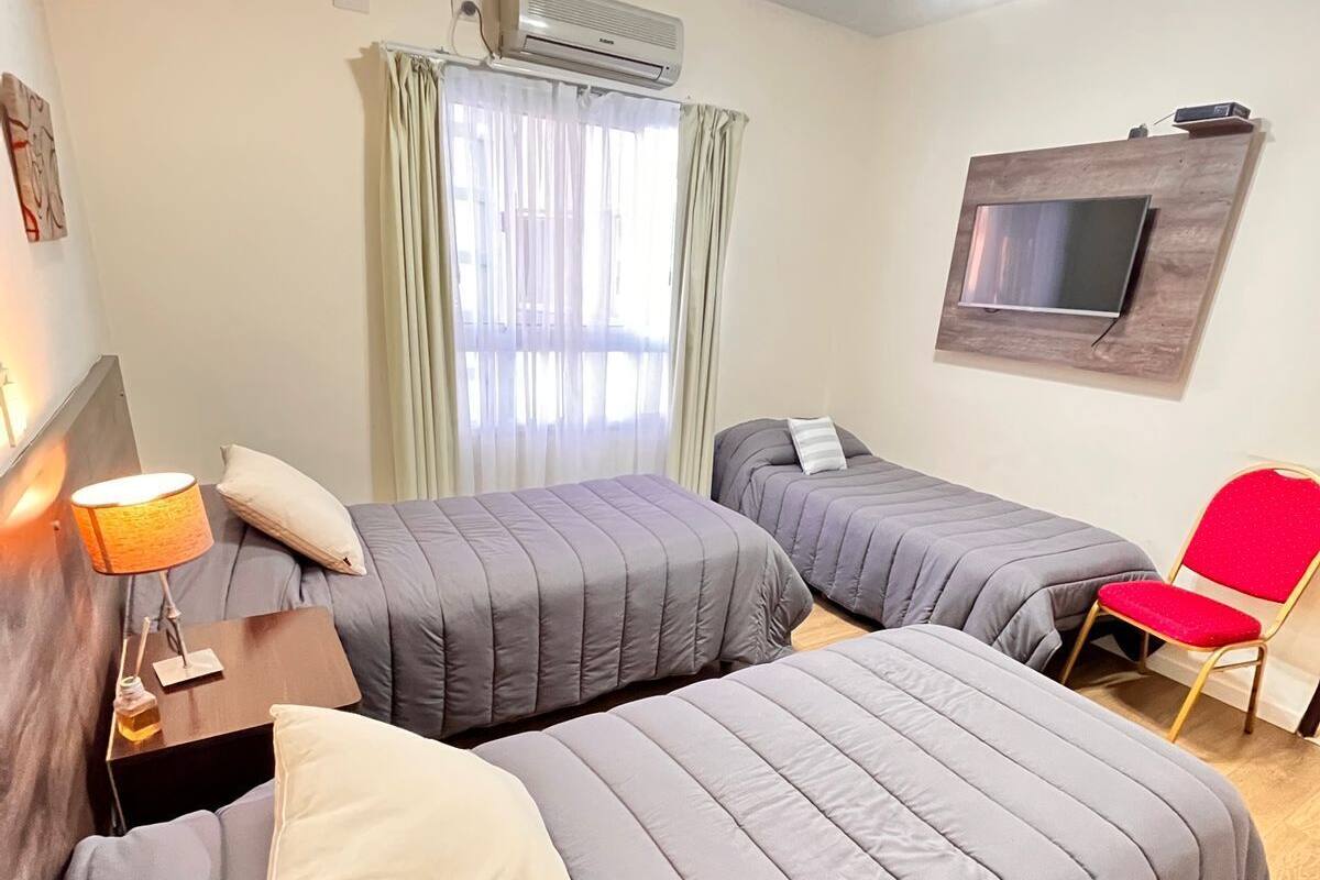 A hotel room with two beds, a TV mounted on the wall, an air conditioner, a ceiling fan, and a red chair.