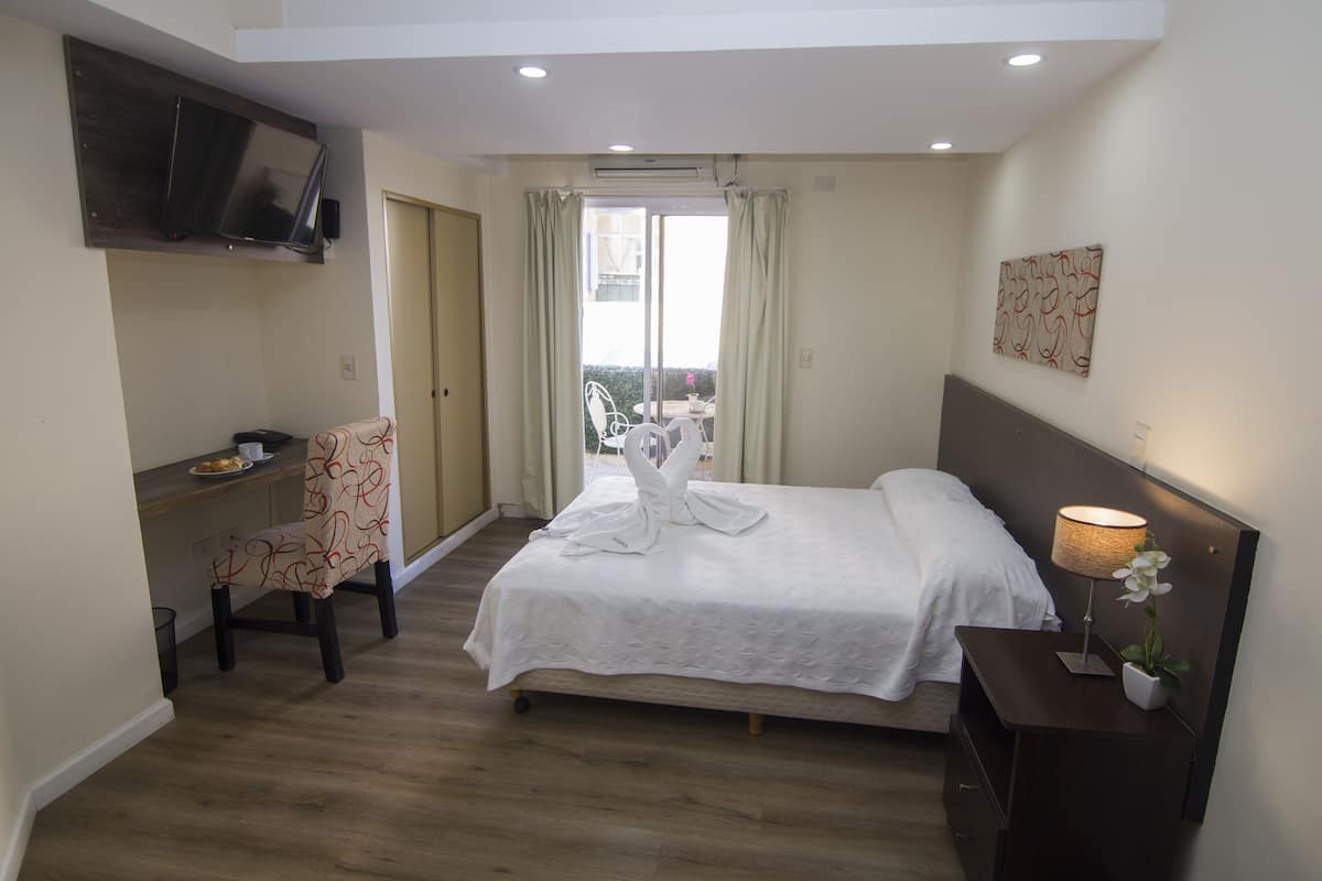 A hotel room with a bed, a desk, a chair, a TV mounted on the wall, and a potted plant.
