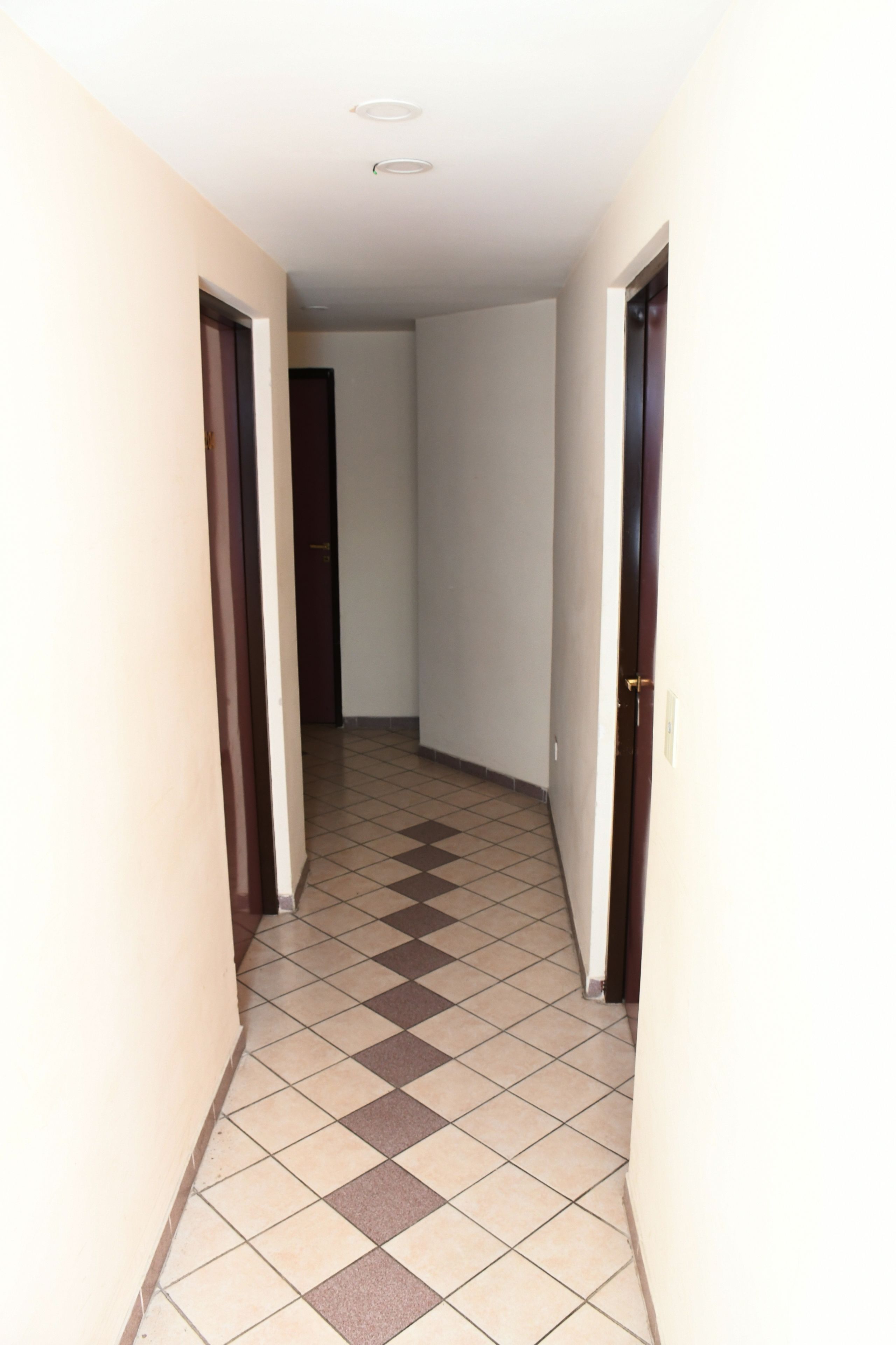 A narrow hallway with checkered floor tiles, white walls, and wooden doors.