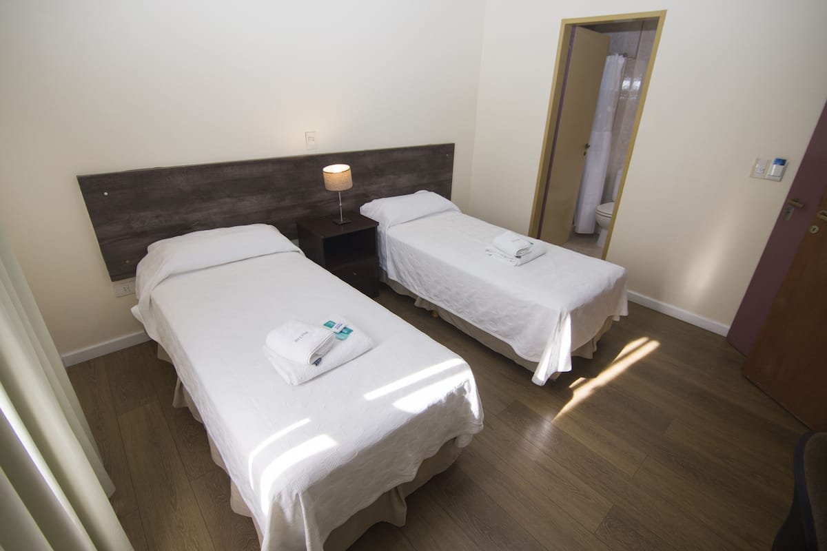 A hotel room with two single beds, a wooden headboard, a nightstand with a lamp, and a mirror on the wall.