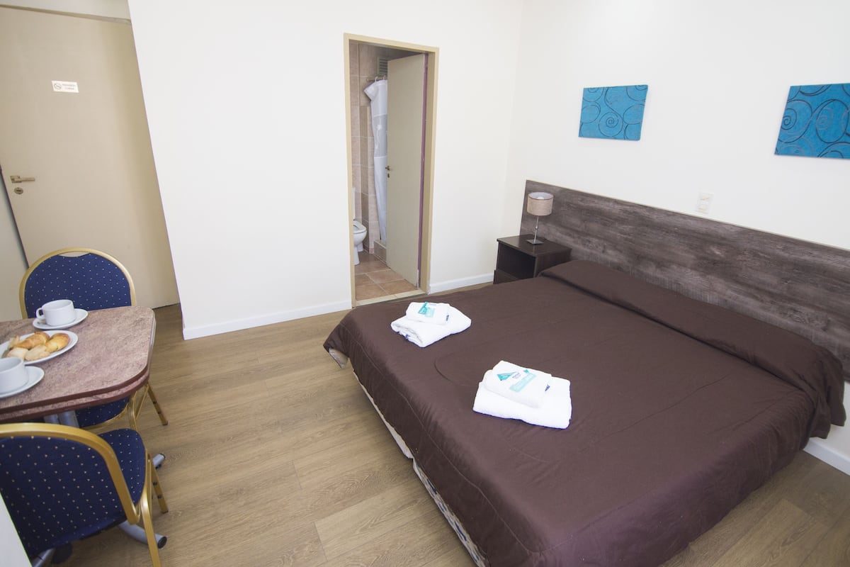 A hotel room with a bed, a small table with a chair, a bathroom visible through an open door, and two wall art pieces.