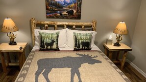 Uma cama com desenho de alce, duas mesas de cabeceira com abajures e um quadro na parede.