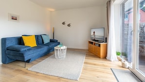 A modern living room with a blue sofa, a small white side table, and a television on a wooden cabinet. - Apartment 1 Küstenkind (ID 391) (Sankt Peter-Ording)