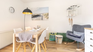 A modern living room with a wooden dining table, white chairs, a grey sofa, a rocking chair, and a painting of a beach scene. - Apartment 1 Küstenkind (ID 391) (Sankt Peter-Ording)