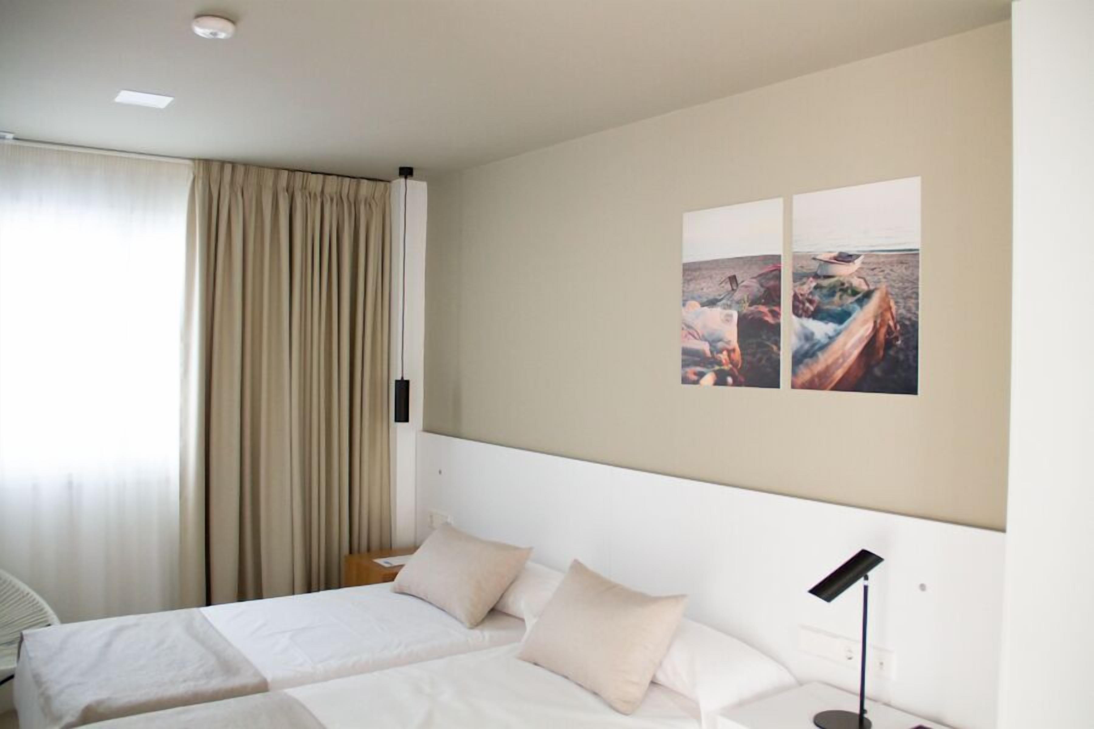 A hotel room with two beds, a large window with curtains, a wall-mounted artwork, and a bedside table with a lamp.