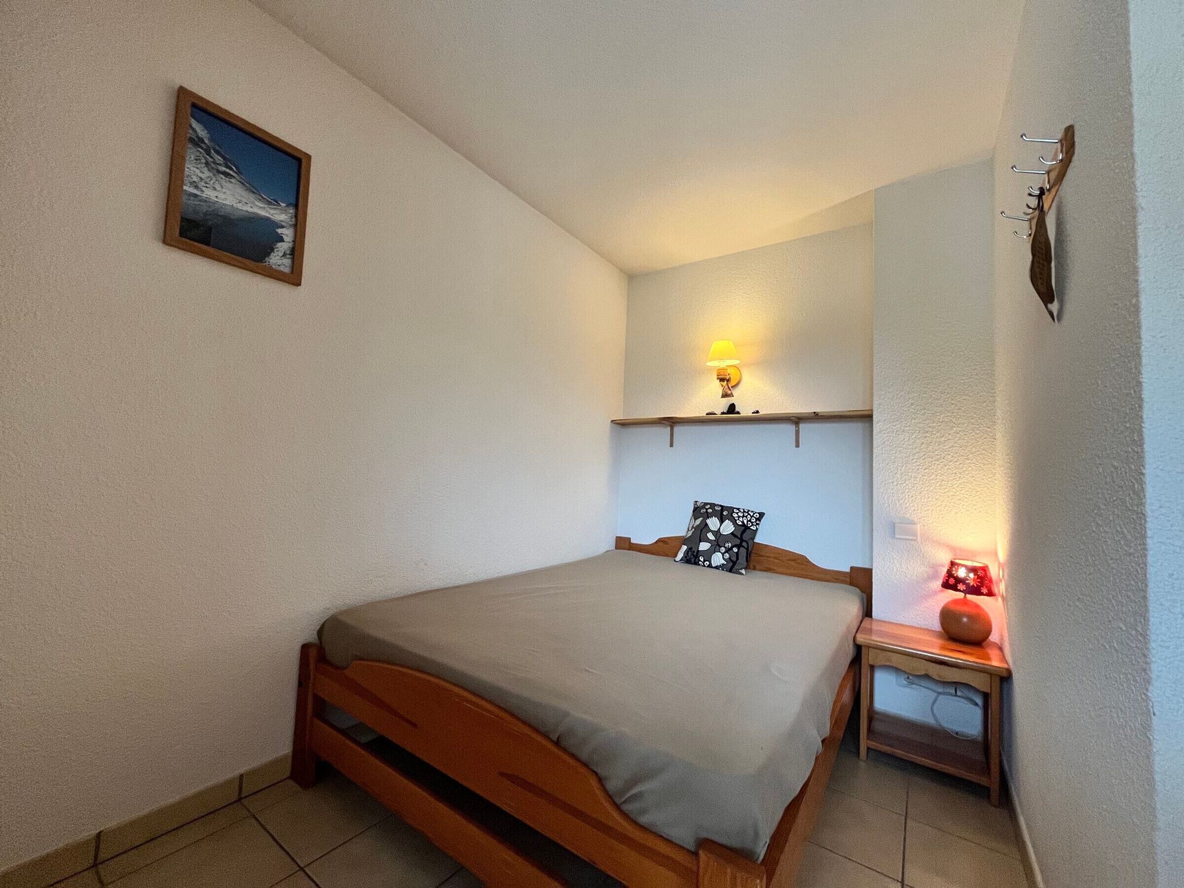 A bedroom with a wooden bed, a nightstand with a lamp, a wall-mounted shelf, and a framed picture.