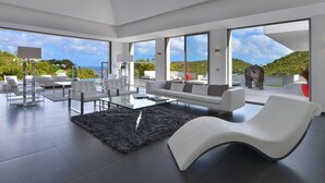A spacious living area with a modern white sofa, a glass coffee table, and a black furry rug. Large windows offer a view of the outdoors.