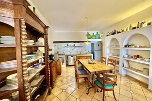 Una cocina con una mesa y sillas de madera, suelo de baldosas y estantes con vajilla y botellas.