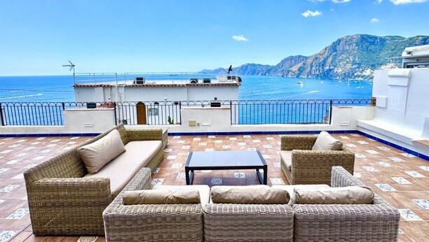 Eine Terrasse mit Korbmöbeln, einem Tisch und Blick auf das Meer und die Berge.