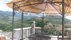 Une terrasse avec une table et des chaises en métal, un parasol en chaume et une vue sur des collines ondoyantes.
