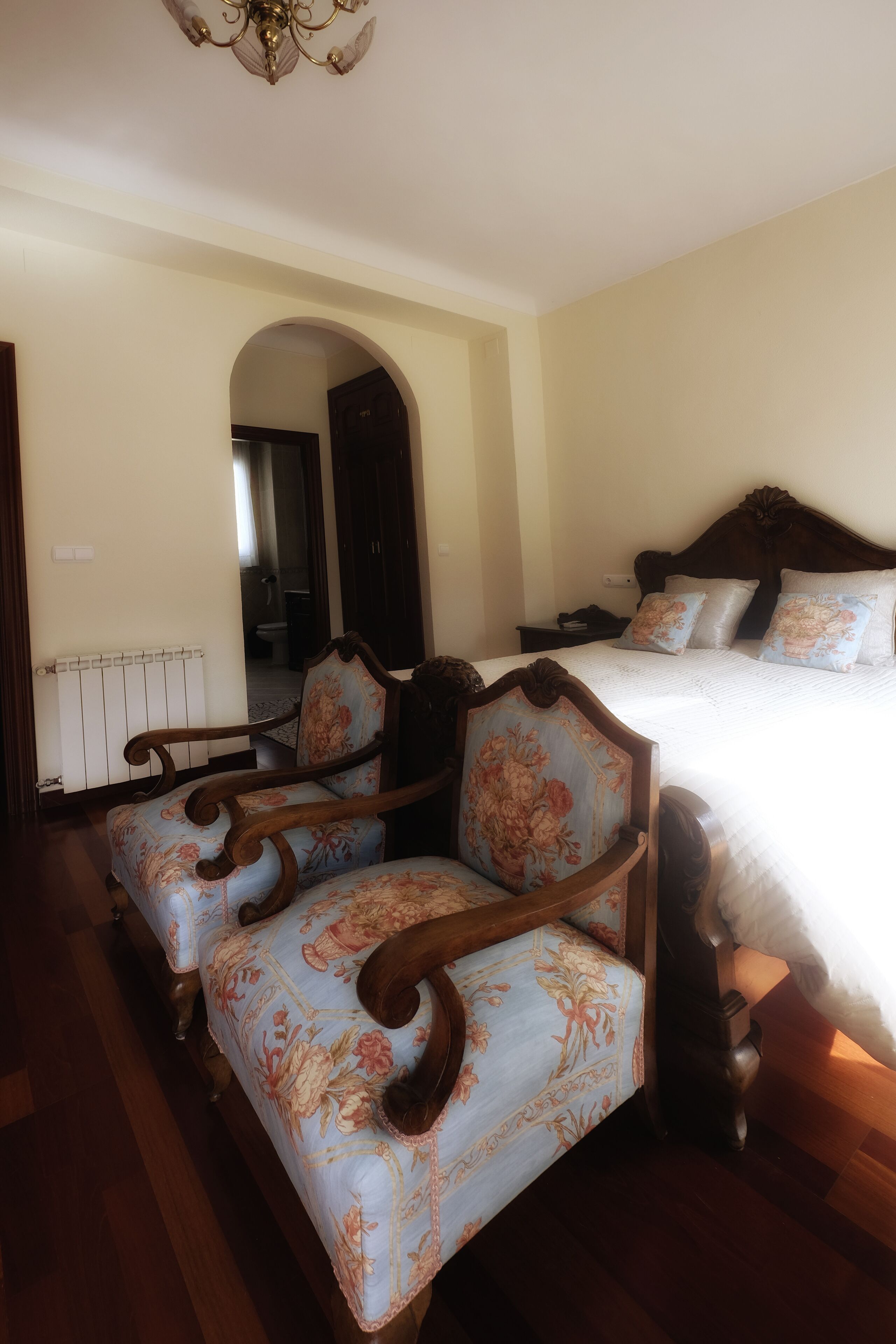 Un dormitorio con una cama grande, dos sillones tapizados, piso de madera, un radiador y un arco que da a otra habitación.