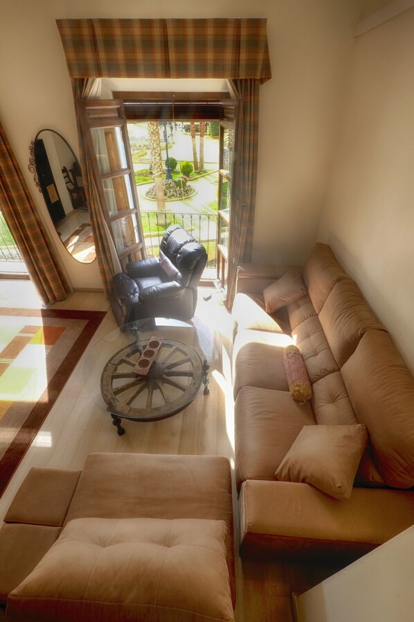 A living room with a sofa, armchair, and ottoman, a round coffee table, and a view of a garden through a window.
