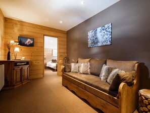 Una habitación acogedora con suelo de madera, un sofá marrón con almohadones mullidos, un escritorio con lámparas y una cama al fondo.