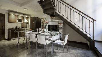 Una cocina moderna con electrodomésticos de acero inoxidable, un comedor con mesa de vidrio y sillas blancas, y una escalera con barandillas de metal.