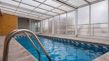 Una piscina interior con paredes de vidrio transparente, una escalera de metal y un borde con azulejos.
