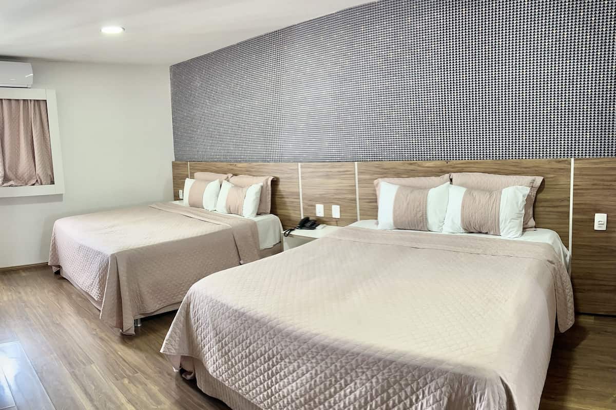 Hotellihuone, jossa on kaksi sänkyä, puinen pääty, seinään asennettu ilmastointilaite ja ikkuna, jossa on verhot.