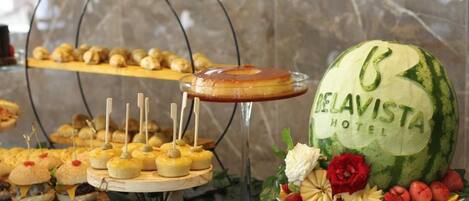 Un bufé con diversos alimentos, incluyendo una sandía como pieza central con la marca 'Delavista Hotel'.