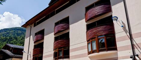 A multi-story building with balconies, a white facade, and a mountainous backdrop.