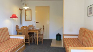 Una habitación pequeña y acogedora con dos camas individuales, una mesa de comedor de madera con sillas, una lámpara roja, un espejo y un cesto de basura verde.
