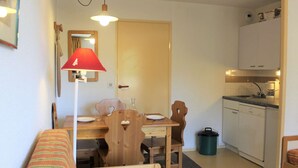 Cocina compacta con armarios blancos, un lavamanos y una cafetera. Hay una mesa de comedor con sillas y una lámpara roja. Un cubo de basura pequeño está cerca del lavamanos.