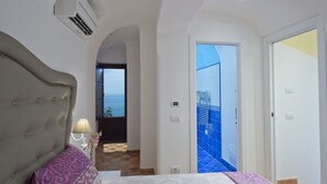 A bedroom with a large bed, a nightstand, and a lamp. There is a view of the sea through a window.
