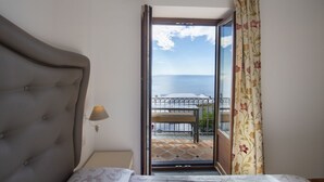 A bedroom with a bed, a nightstand, a lamp, a window with a view of the sea, and a balcony with a railing.
