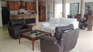 Uma sala de estar ampla com um sofá branco, duas poltronas de couro e uma mesa de centro de vidro. Uma cozinha com armários de madeira e um espaço bar é visível ao fundo.