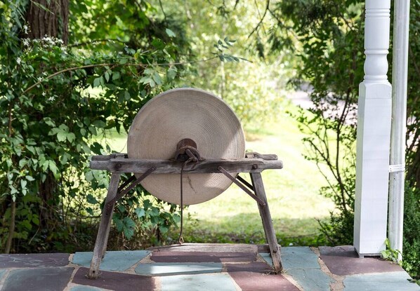 A traditional spinning wheel outdoors, supported by a wooden frame, set on a stone-paved area with a white column nearby.