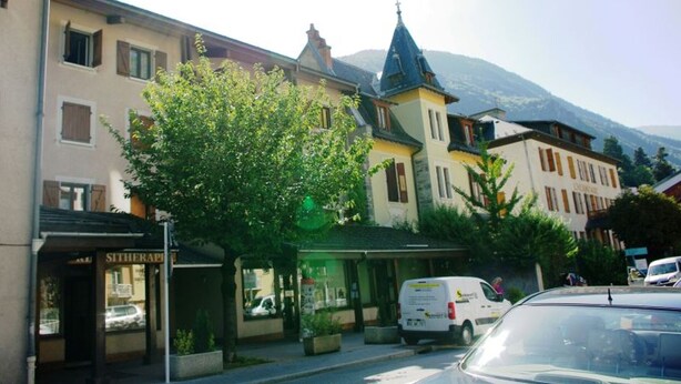 A street view with a row of buildings, including a hotel with a sign that reads "HOTEL". There is a tree in the foreground, and a van is parked on the street. - Central studio with parking, 2 people. (Brides-les-Bains)