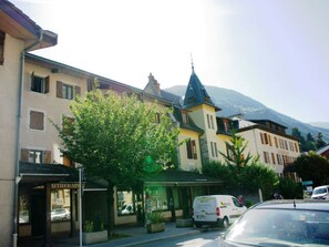 A street view with a row of buildings, including a hotel with a sign that reads "HOTEL". There is a tree in the foreground, and a van is parked on the street. - Central studio with parking, 2 people. (Brides-les-Bains)