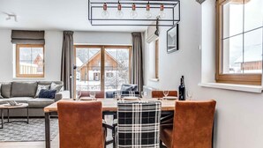 Ein Essbereich mit Tisch für vier Personen, einer Couch und Blick auf eine Holzhütte durch große Fenster.