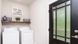 Une buanderie avec deux machines à laver blanches, une étagère en bois contenant des produits de lessive et une porte noire dotée d’une fenêtre en verre dépoli.