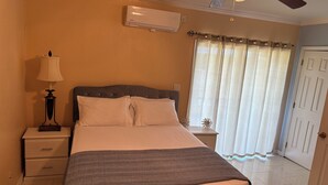 A bedroom with a bed, bedside table, lamp, air conditioning unit, and a window with curtains.