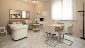 A modern living room with a sofa, a dining table with chairs, a small side table, a large mirror, and a window with curtains.