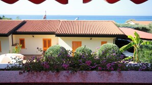 Un edificio costero con techo de tejas, un jardín con flores moradas y vista al mar.