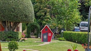Un jardín con una casita de juegos roja, un árbol grande y un sube y baja rojo y amarillo.