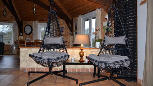 A cozy room with a wooden ceiling, a hanging chair with cushions, a table with a lamp, and a clock on the wall. - Gîte le Manège normand (Grand-Camp)