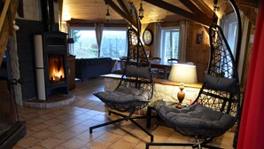 A cozy room with a wood-burning stove, two cushioned chairs, a dining table, and a view of the outdoors. - Gîte le Manège normand (Grand-Camp)