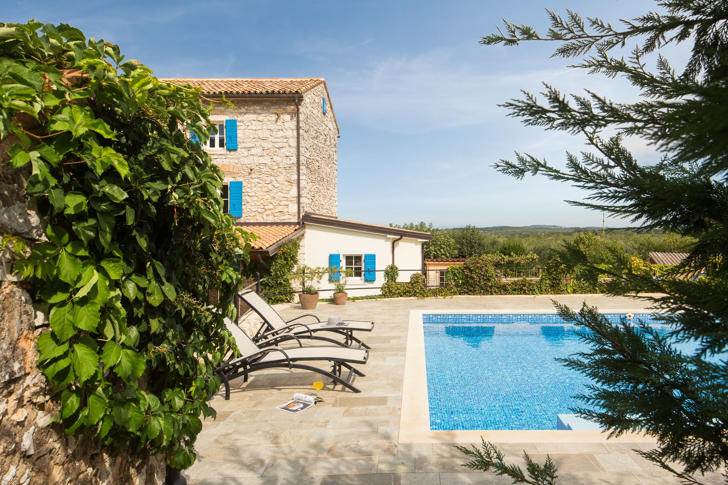A stone building with blue shutters, a swimming pool, and a garden with a tree.